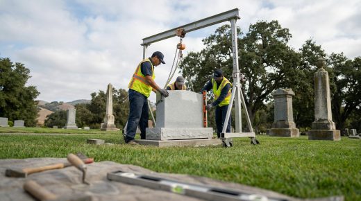 Trois ouvriers en gilets de sécurité installent une stèle en granit dans un cimetière à l'aide d'un portique de levage.