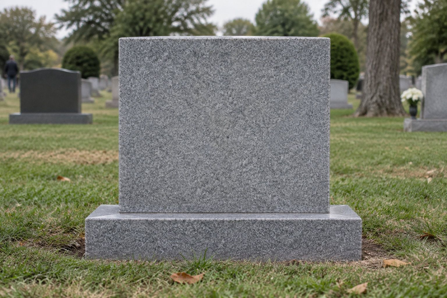 Une grande stèle funéraire grise sans inscription se dresse dans un cimetière herbeux, entourée d’autres pierres tombales, d’arbres et de fleurs.